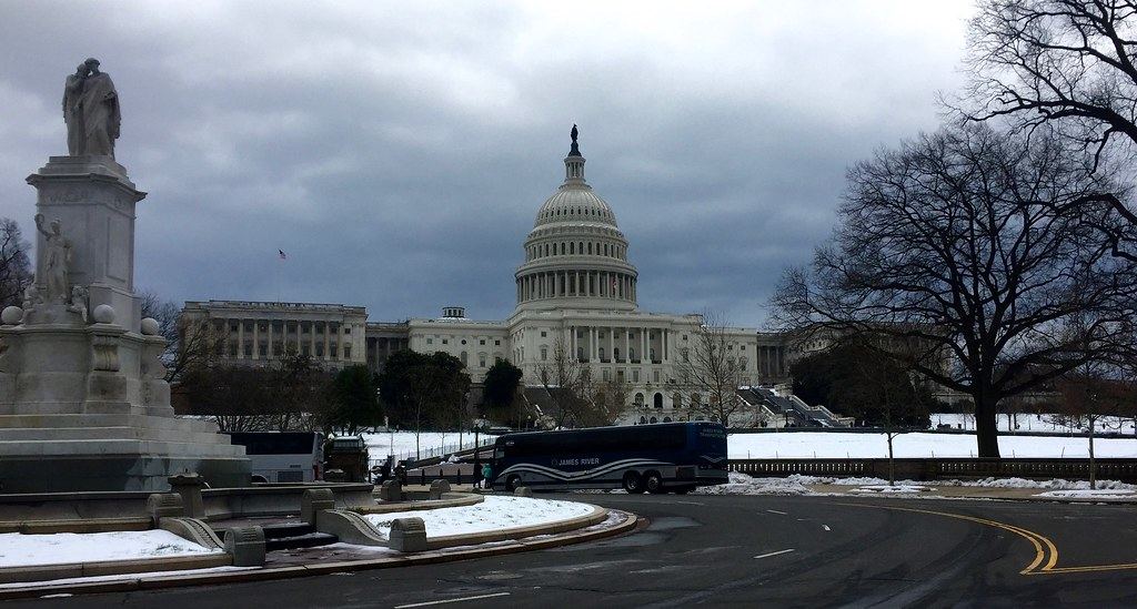 The U.S. Capitol dome under cloudy skies, with snow on the grounds, a tour bus and a monument in front