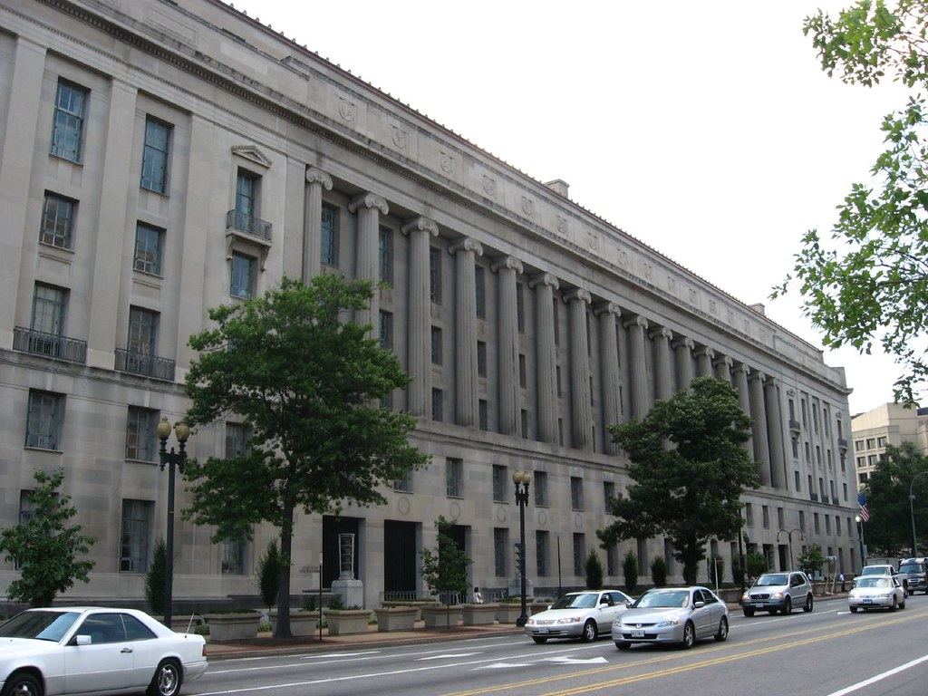 Large neoclassical stone government building with tall columns along a tree-lined street with passing cars