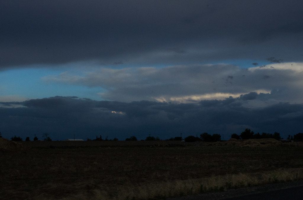 A dark, flat farm field stretches beneath heavy storm clouds at dusk, with a line of trees on the horizon