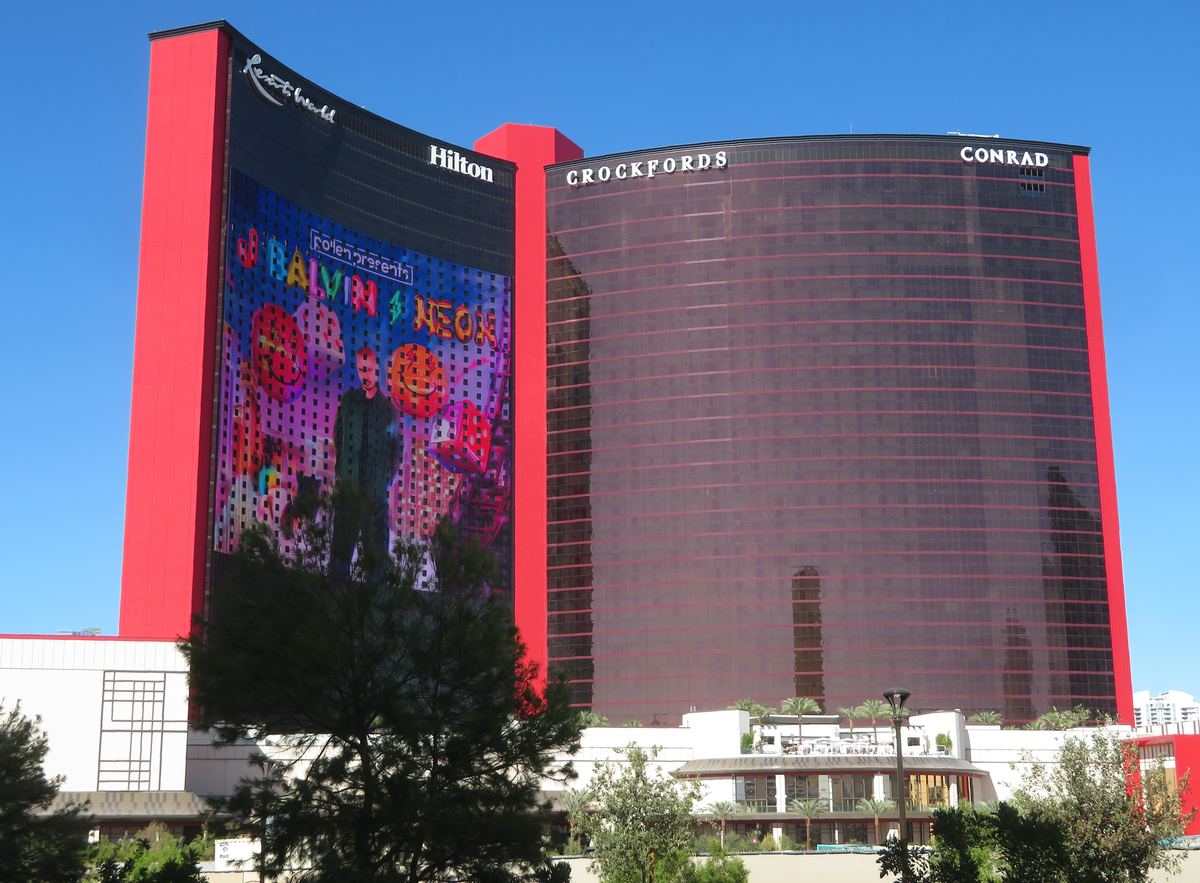 Curved red-trimmed towers of Resorts World Las Vegas with a large LED advertising screen under a clear blue sky