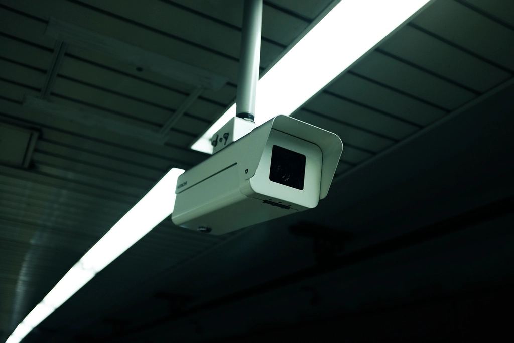 A white security camera mounted on a pole hangs from a dark ceiling beside a long fluorescent light