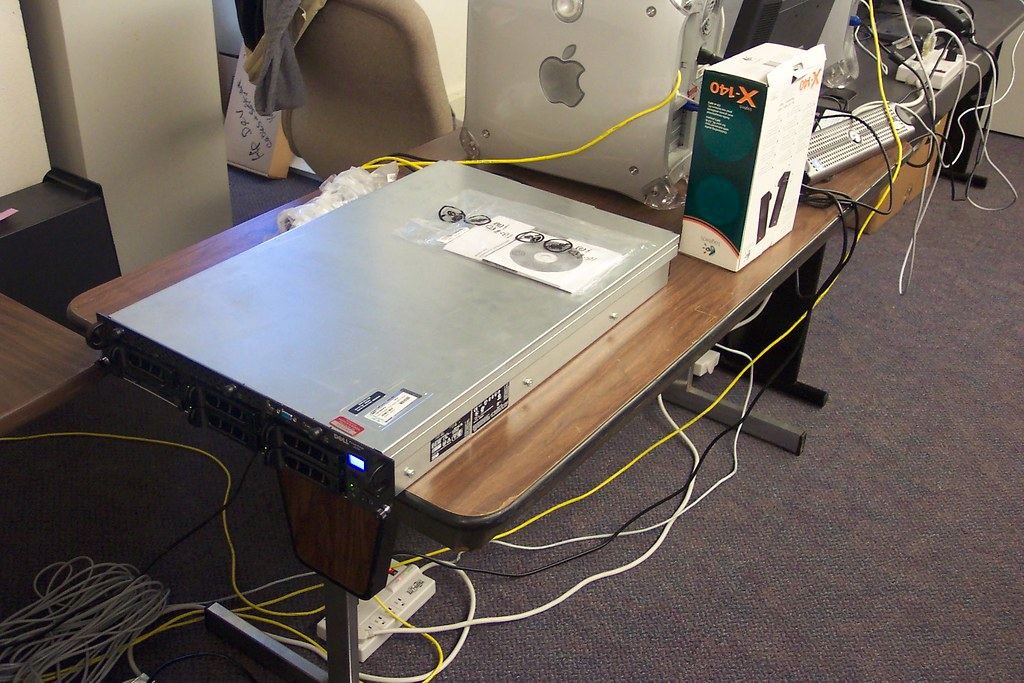 A rack-mount server sits on a wooden office table surrounded by cables, a power strip and a desktop computer