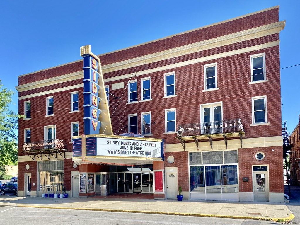 Red brick theater building with a tall vertical 'Sidney' sign and a lettered marquee over the entrance