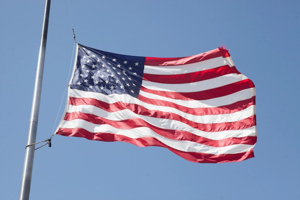 An American flag with a frayed edge waves from a metal flagpole against a clear blue sky
