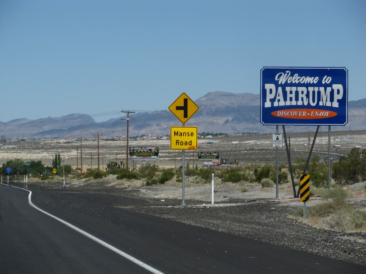 A blue Welcome to Pahrump sign and a Manse Road sign beside a desert highway with mountains behind
