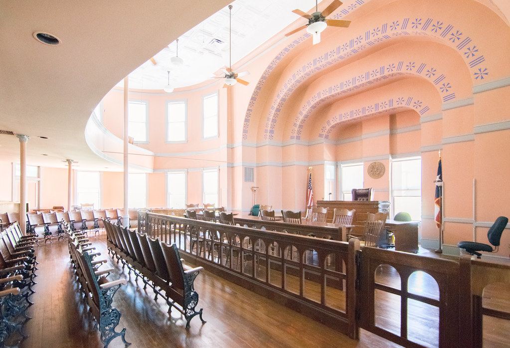 An empty historic courtroom with peach walls, arched ceiling, wooden spectator seats and a raised judge's bench