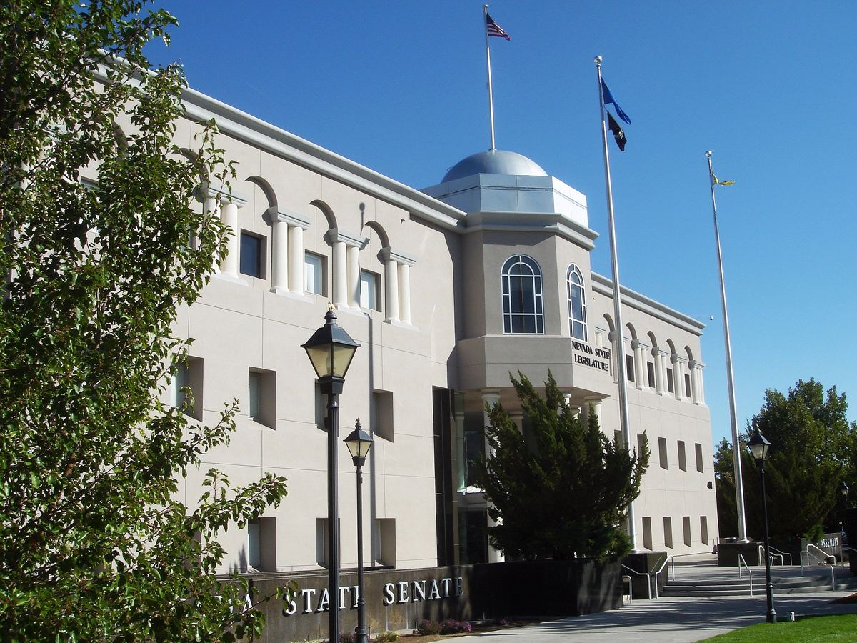 A beige domed government building with arched windows, flagpoles and lampposts under a clear blue sky