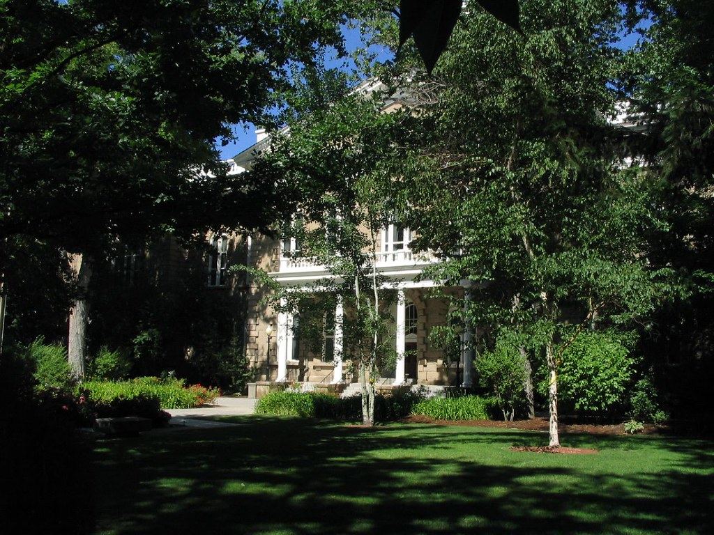 A sandstone building with white columns and a portico, partly hidden by leafy trees beyond a shaded green lawn
