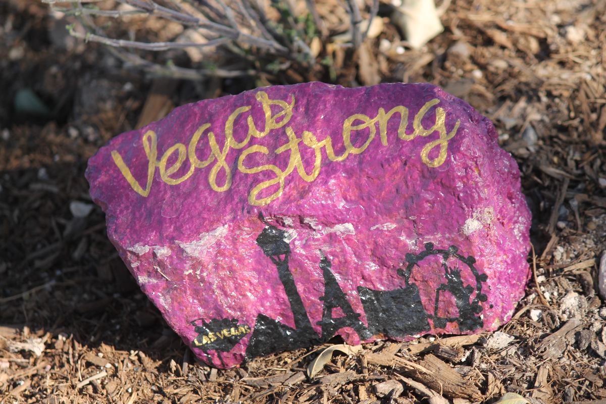 A pink painted rock reading 'Vegas Strong' with a black Las Vegas skyline silhouette, resting on wood mulch