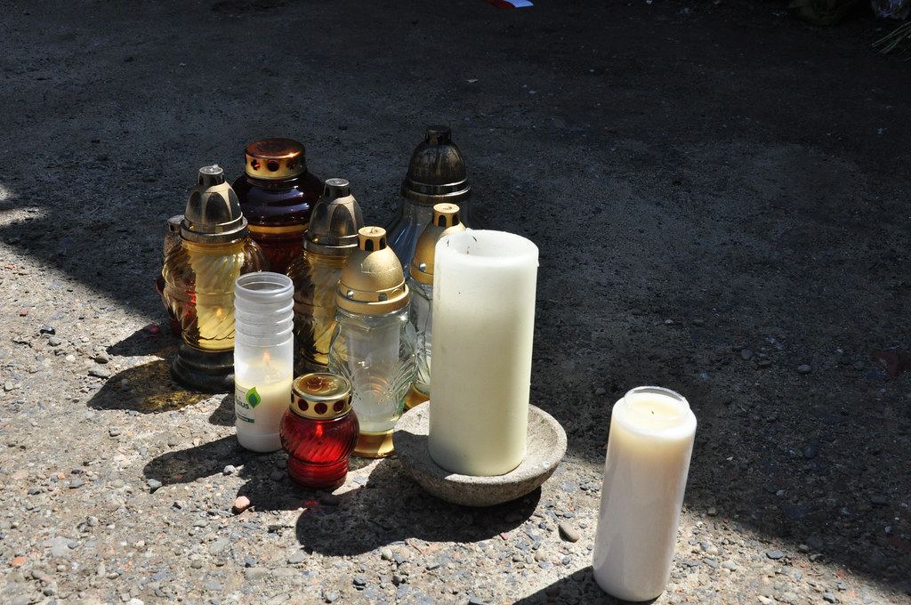 A cluster of memorial candles and glass grave lanterns sits on sunlit gravel ground
