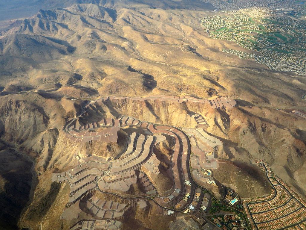 Aerial view of terraced home sites and winding roads carved into desert hills beside a suburban neighborhood
