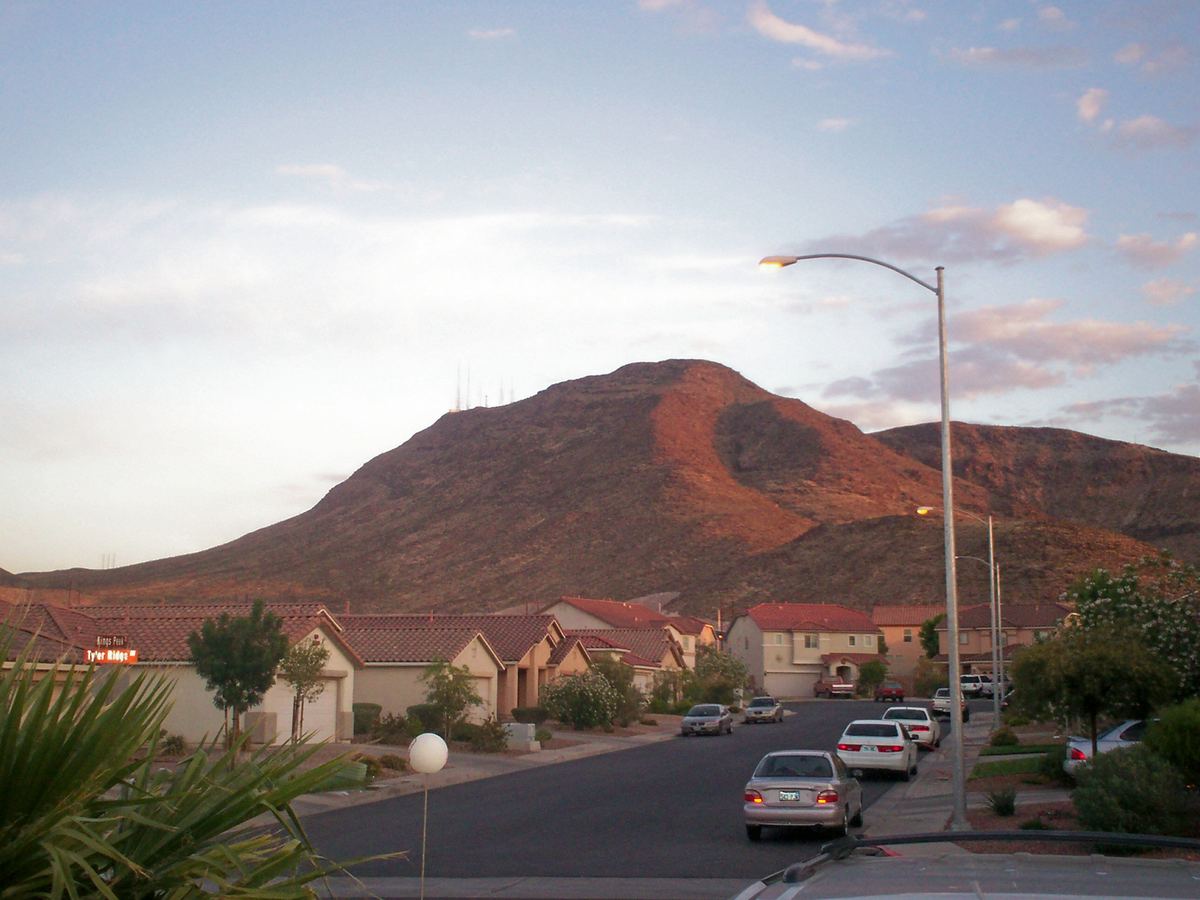 Rocky desert mountain with antennas on top rises behind a street of tile-roofed homes and parked cars at dusk