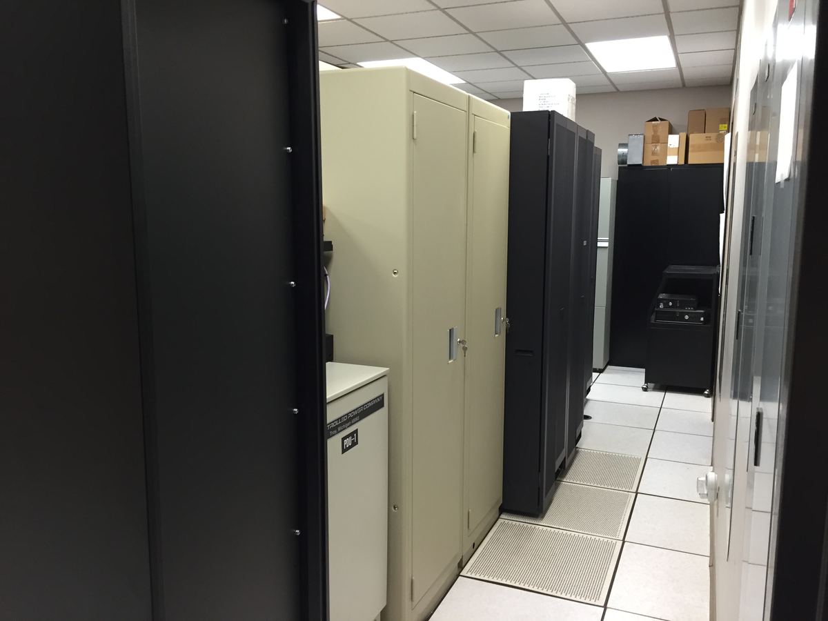 Narrow server room with black and beige equipment cabinets, a raised tile floor and boxes stacked near the ceiling