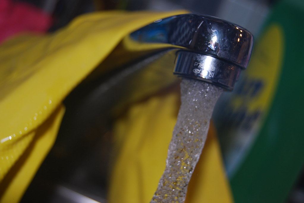 Close-up of a chrome kitchen faucet with water flowing, yellow rubber gloves blurred in the background