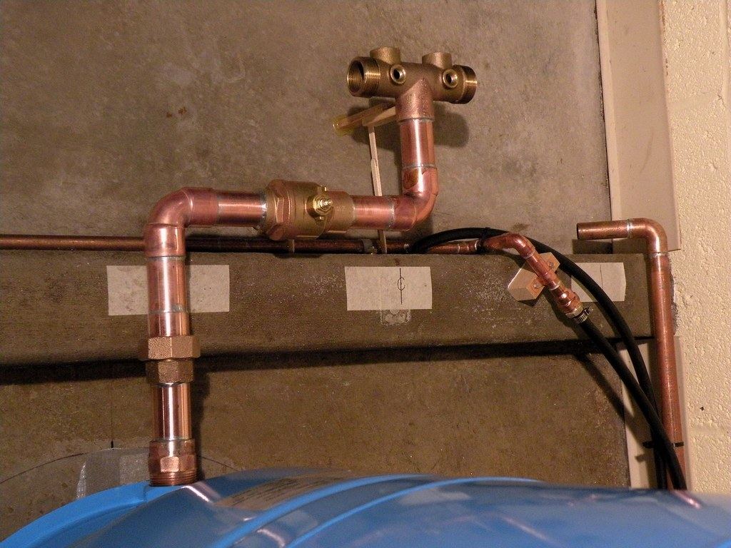 Copper water pipes with a brass ball valve and fittings above a blue pressure tank against a concrete wall