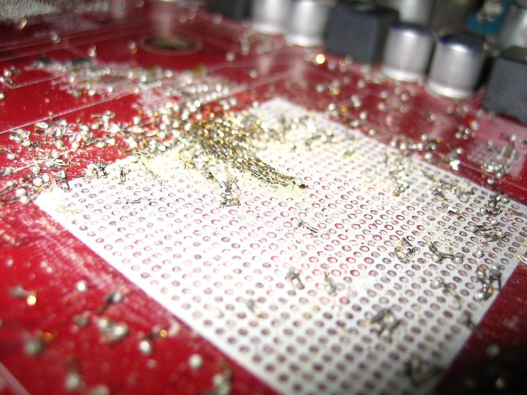 Close-up of a red circuit board with a white chip socket area scattered with bits of solder and small components