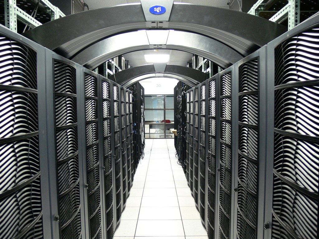 Rows of black server racks with curved grilles line a white-tiled aisle under arched ceiling panels
