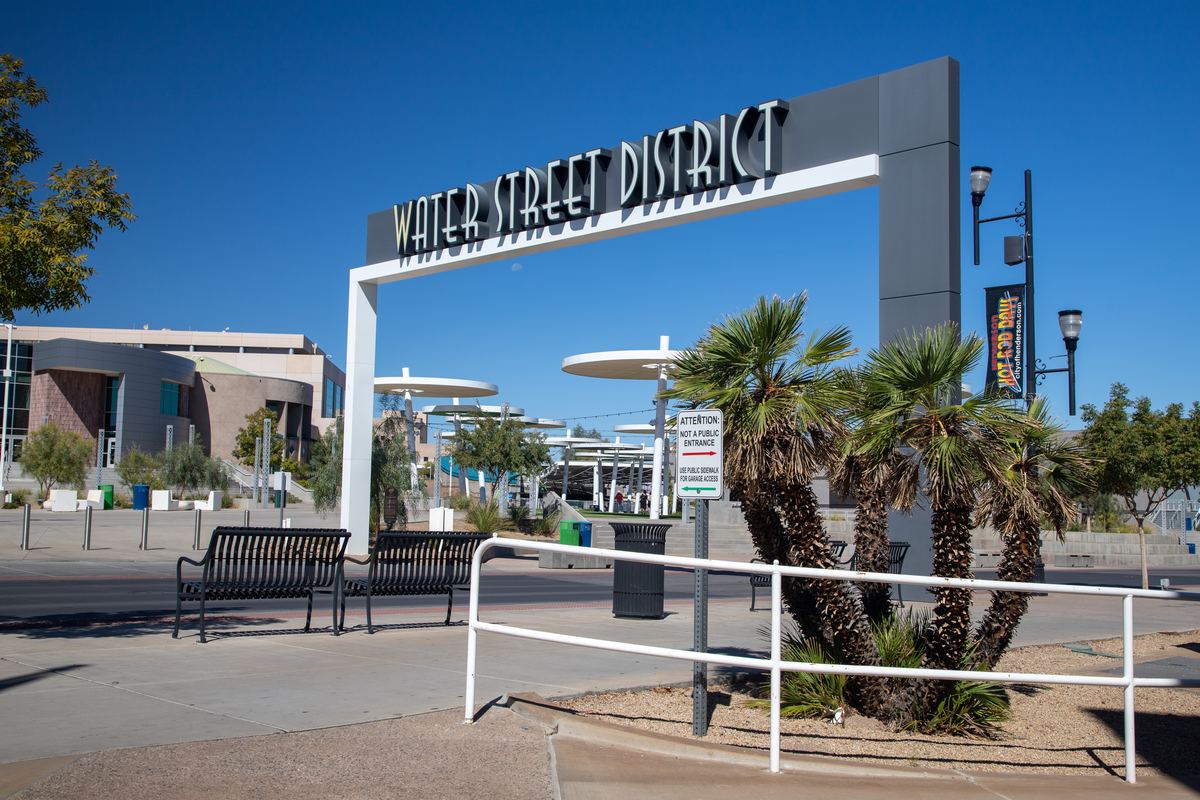 A Water Street District archway sign over a plaza with benches, palm trees and shade canopies under a clear blue sky