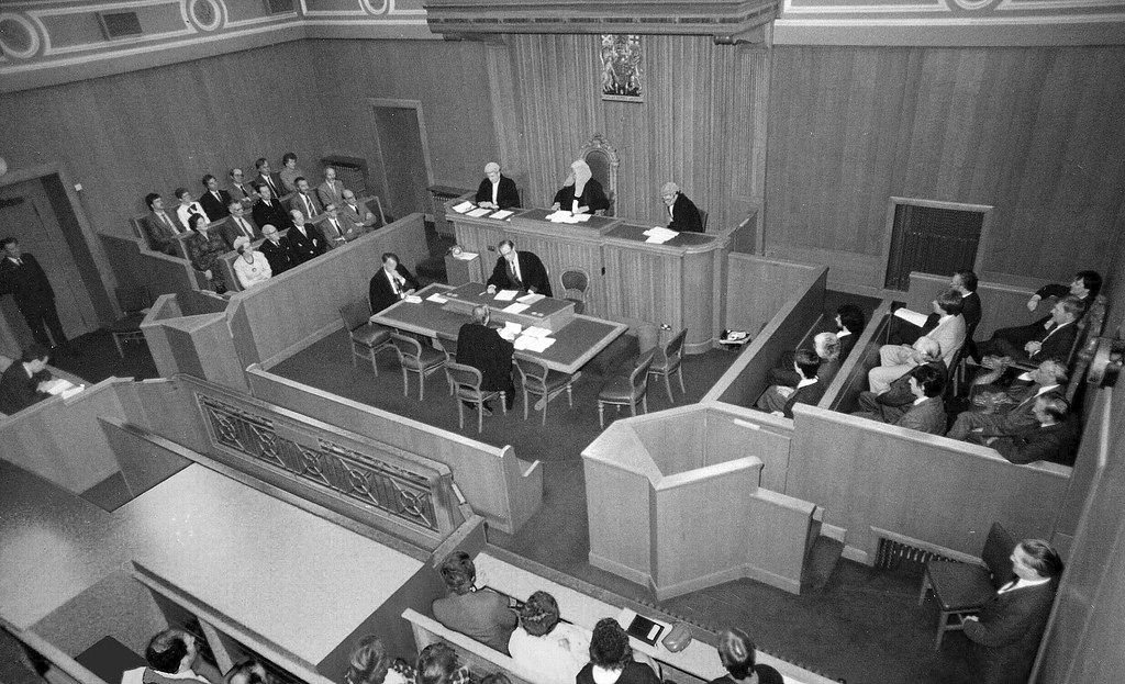 Black-and-white overhead view of a wood-paneled courtroom with judges at a raised bench and people seated in pews