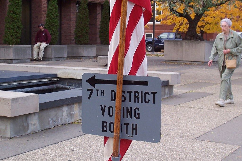 A sign reading '7th District Voting Booth' with an arrow, mounted on a pole beside an American flag on a plaza