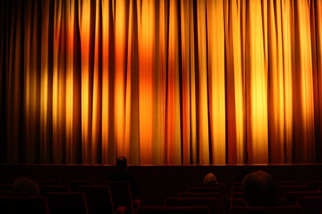 A few people sit in dark theater seats facing a closed orange-lit stage curtain