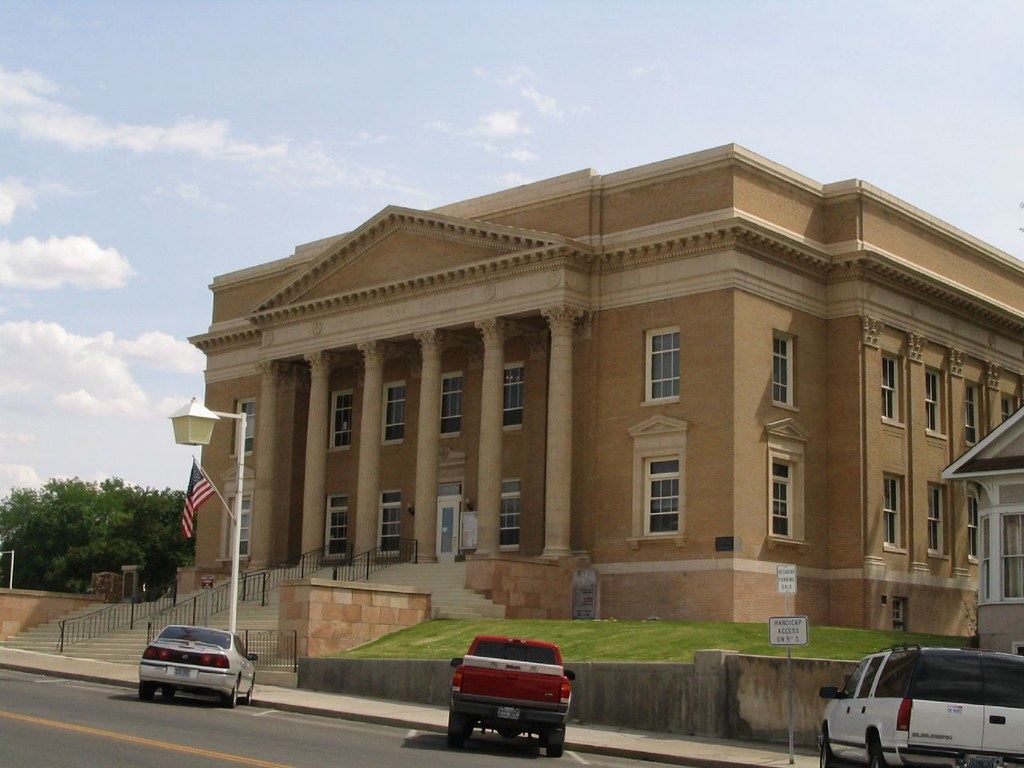 A tan brick neoclassical courthouse with tall columns and wide front steps, with cars parked on the street