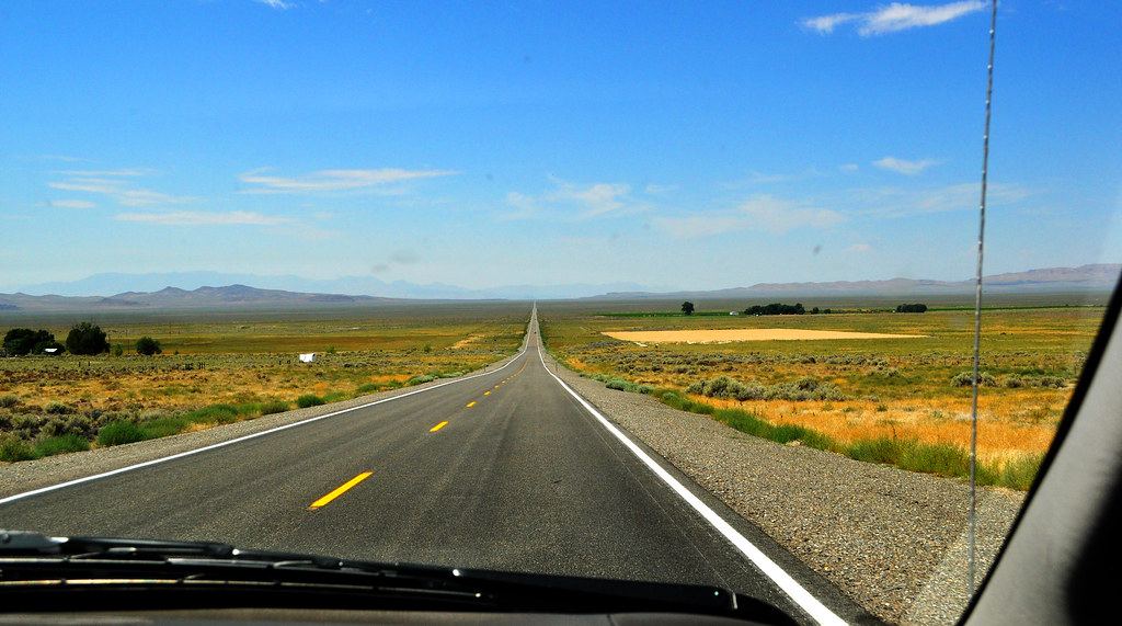 A straight two-lane highway stretches through sagebrush flats toward distant mountains under a blue sky
