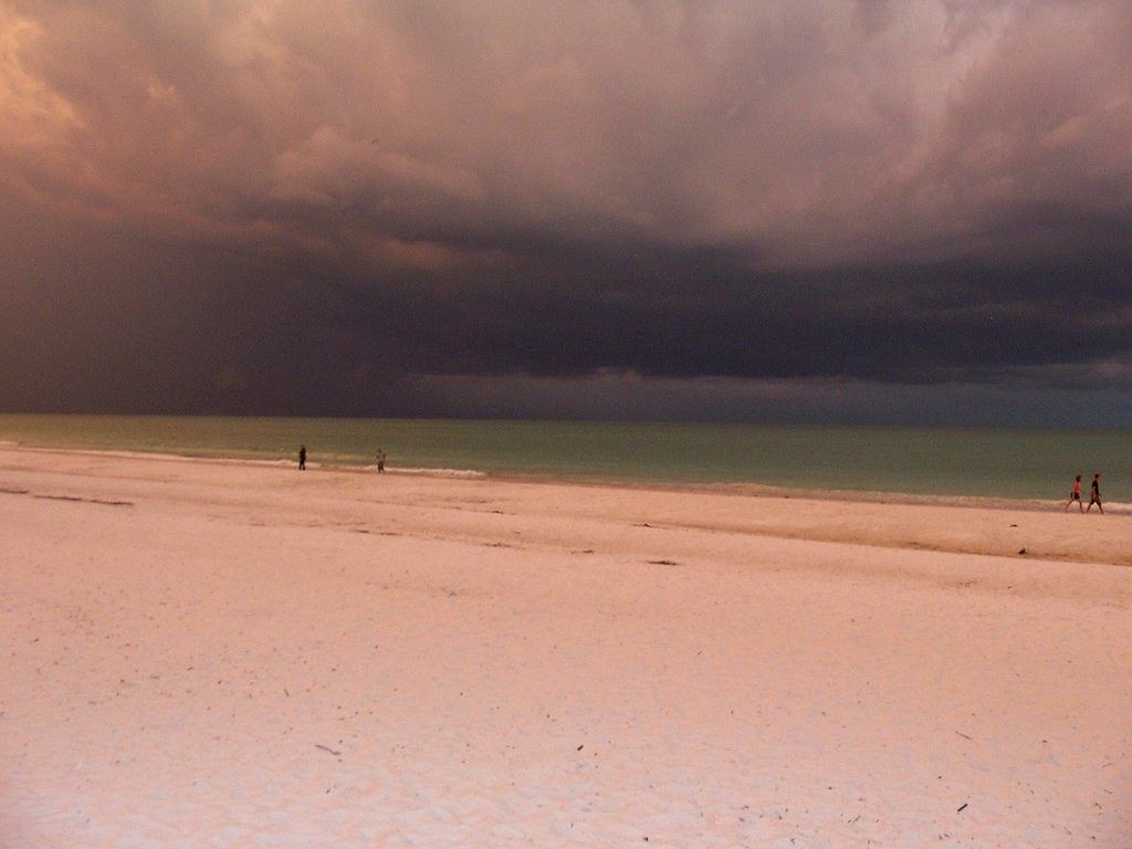 Dark storm clouds hang over a white sand beach and green sea, with a few distant people walking near the water
