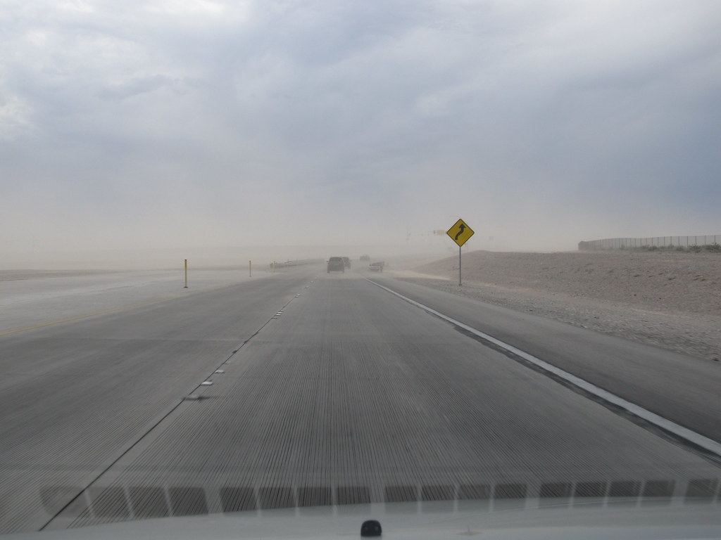 A divided desert highway under hazy, dusty skies with a yellow curve warning sign and distant cars