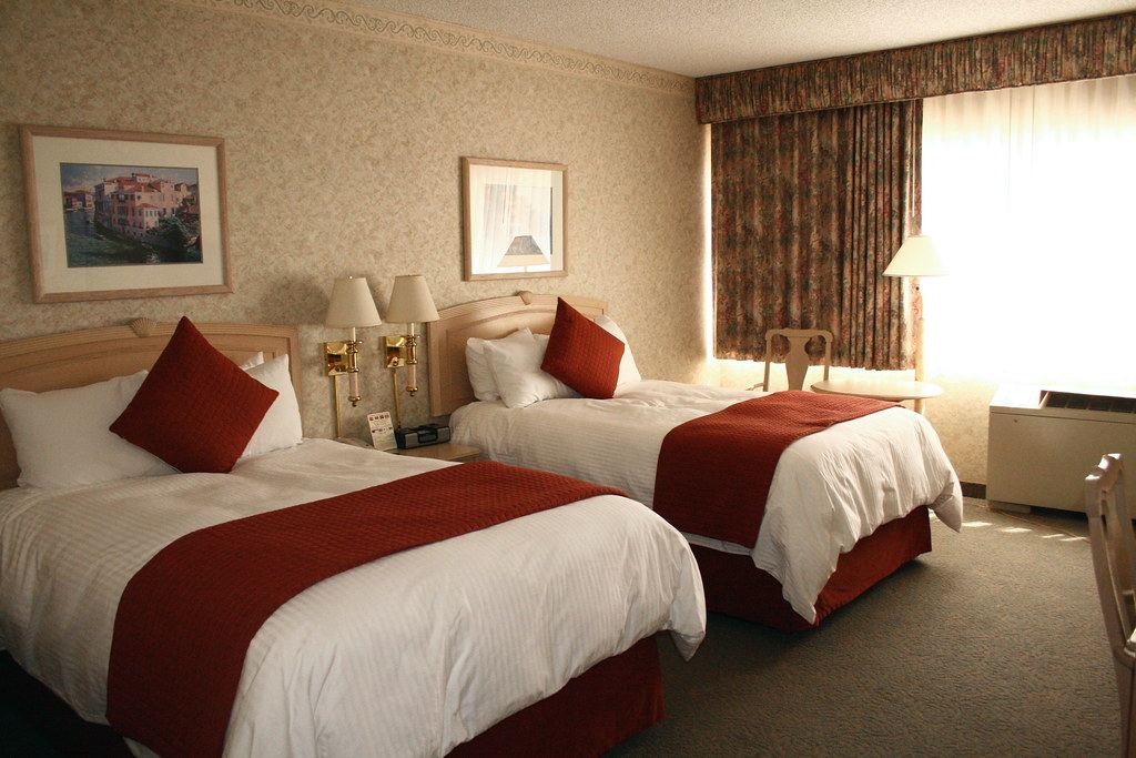 A hotel room with two beds made with white linens and red runners, wall lamps, framed art and floral curtains