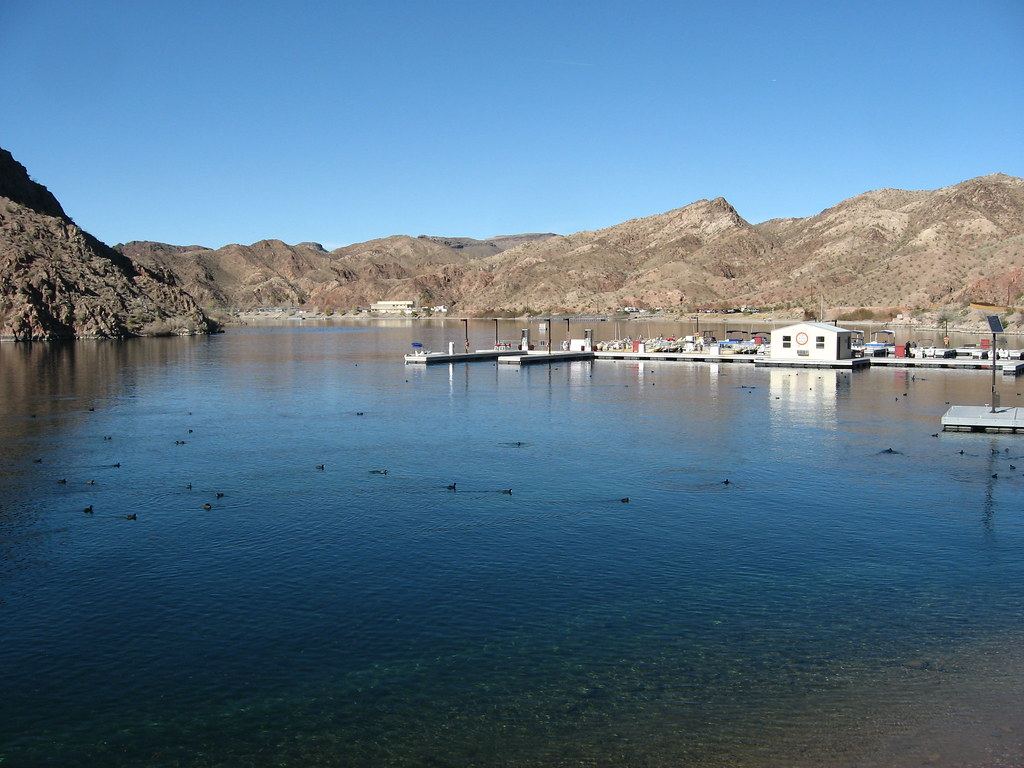 A calm blue lake with a floating marina and docks, set against rugged brown desert hills under a clear sky