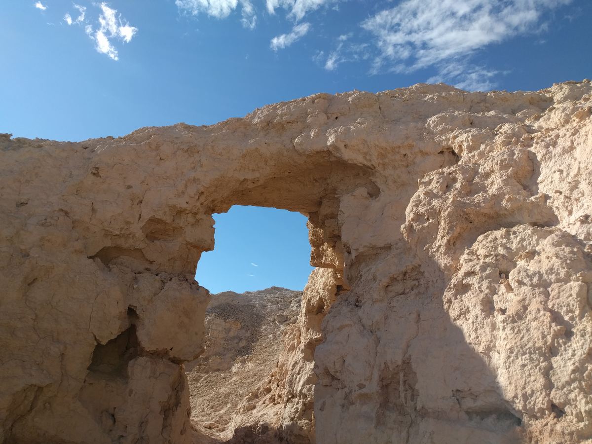 A natural arch in pale, eroded desert rock frames a patch of blue sky under scattered clouds