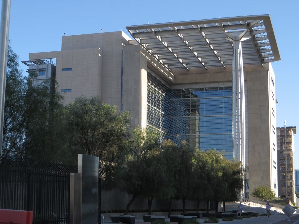 A tall stone and glass courthouse with a large overhanging canopy roof, behind trees and a black iron fence