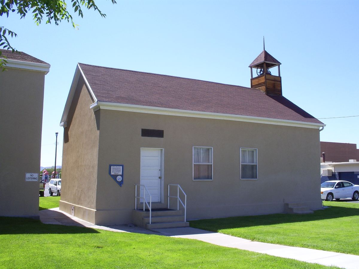 A small tan stucco chapel with a brown shingle roof and wooden bell tower on a sunny lawn