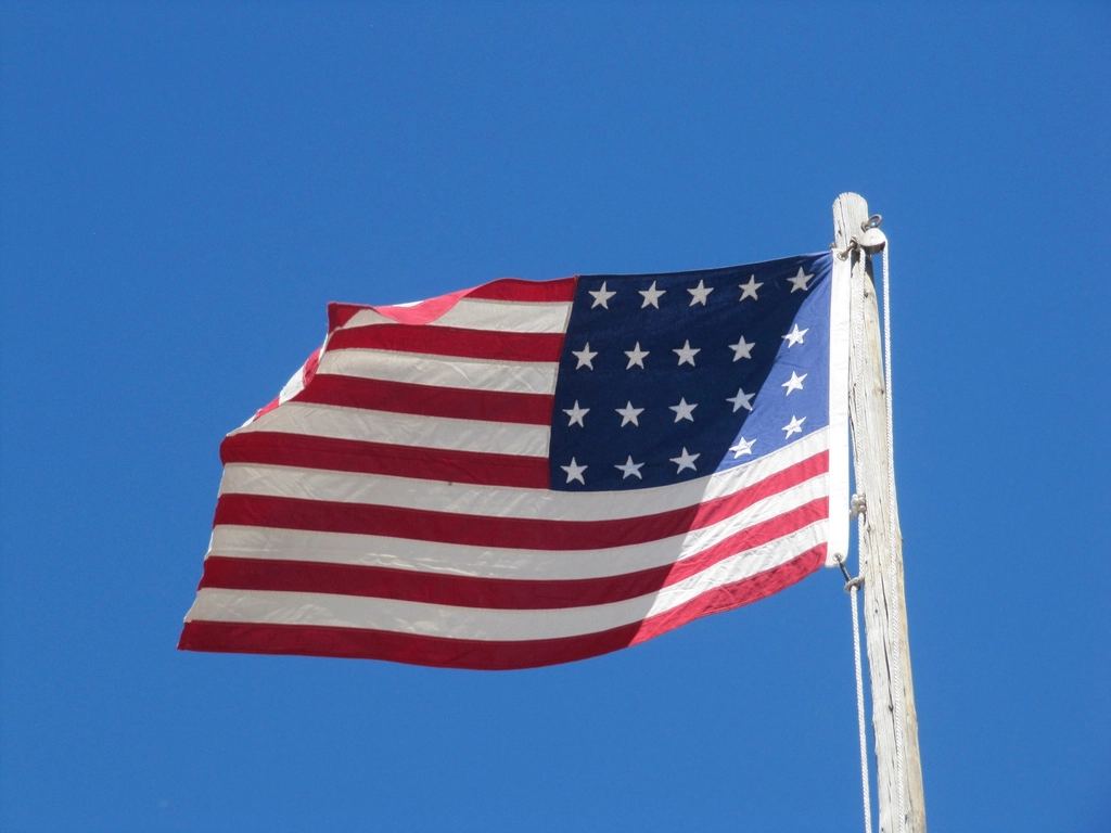 A historic-style American flag with stars and stripes waves from a wooden pole against a clear blue sky