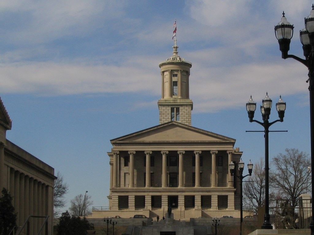 A columned stone government building with a tall cupola and flag under a partly cloudy sky, flanked by lampposts