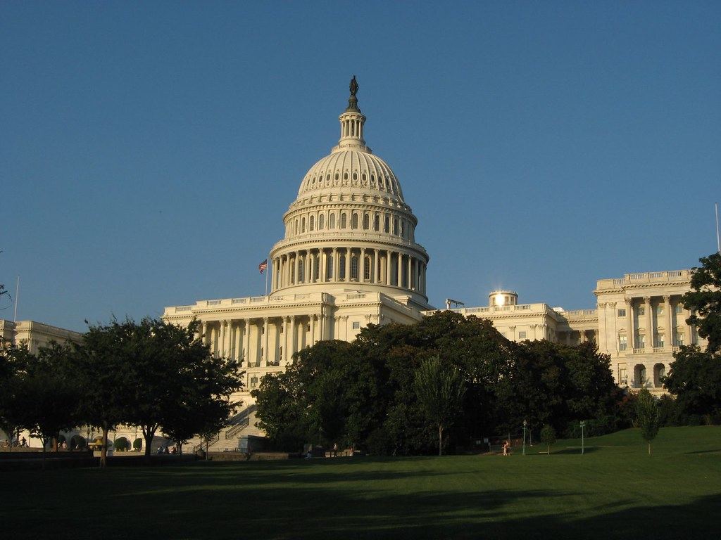 The white domed U.S. Capitol building behind green trees and a lawn under a clear blue sky