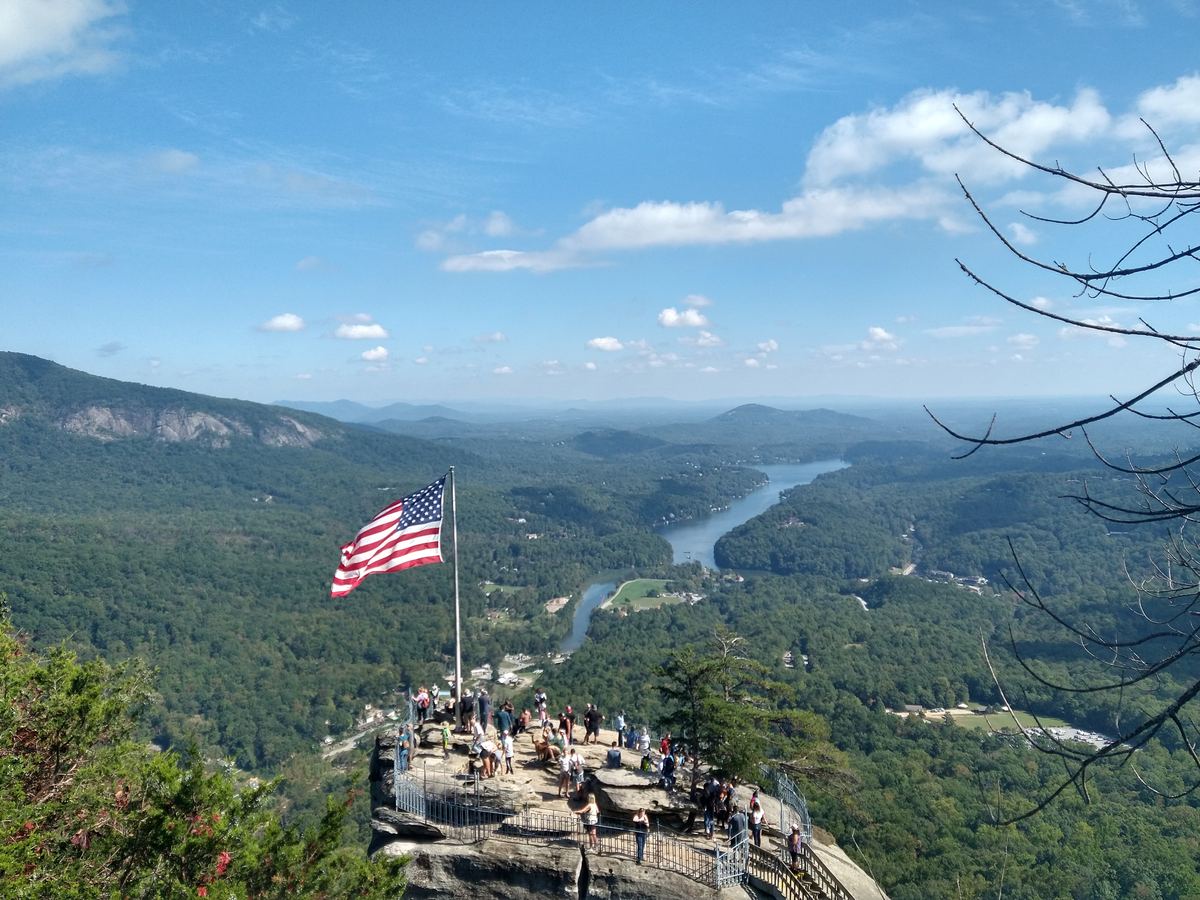 An American flag waves over a rocky overlook with visitors, above a forested valley and a winding lake