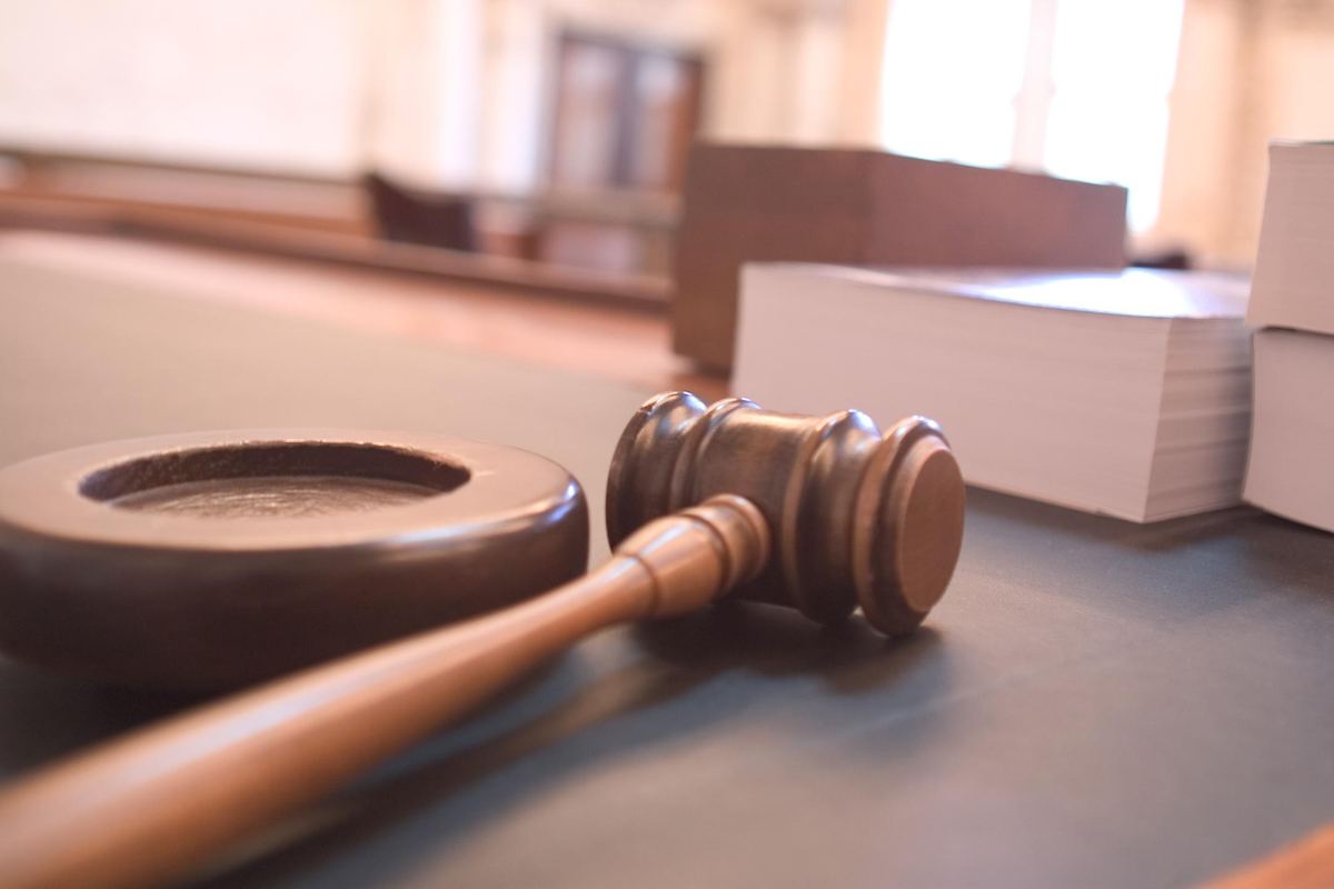 A wooden gavel and sound block rest on a table beside stacks of books in a softly lit courtroom