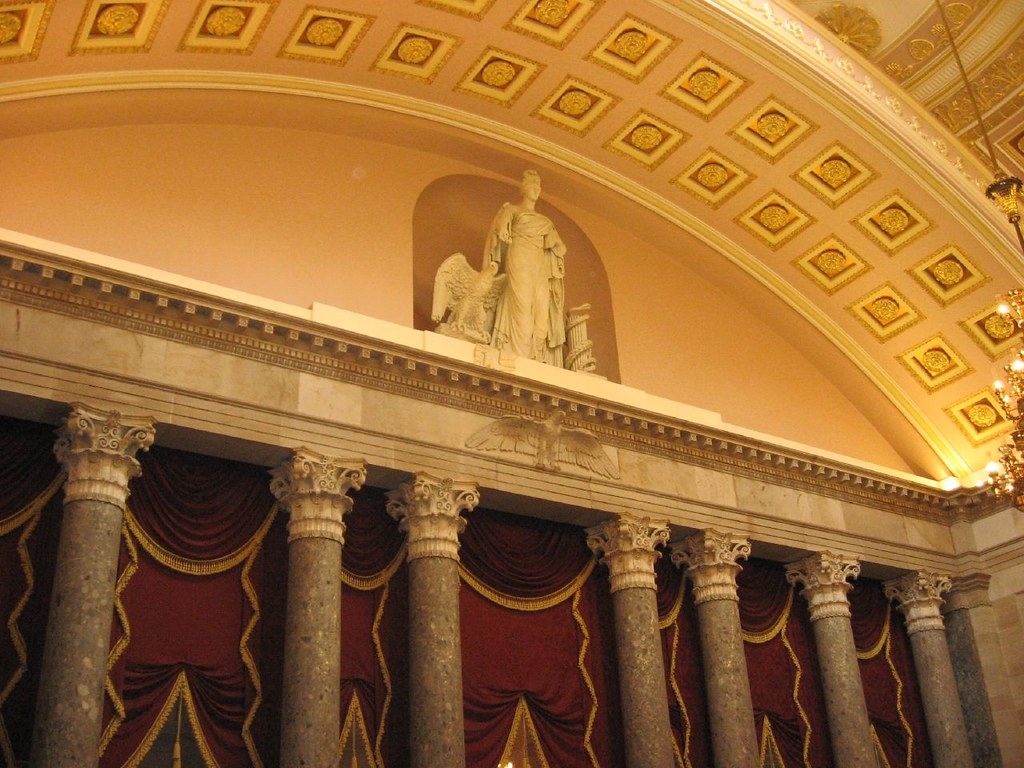 Ornate hall with a coffered gilded ceiling, a marble statue in a niche, and stone columns before red drapes