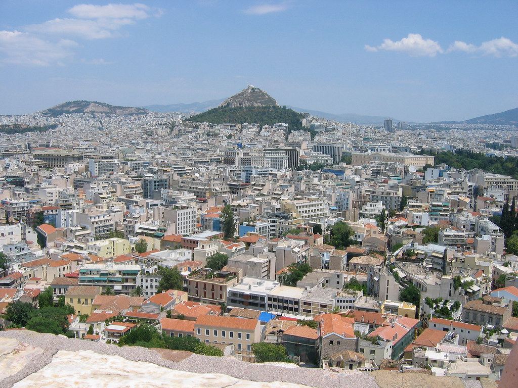 Dense white buildings of Athens spread toward a tree-covered hill under a blue sky