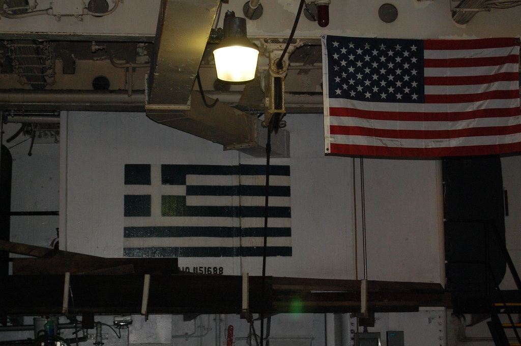 A Greek flag painted on a white wall beside a hanging American flag under an industrial lamp