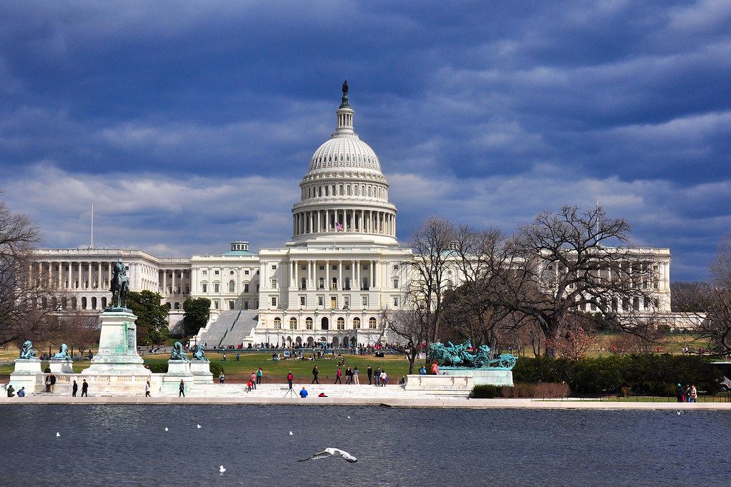 The US Capitol dome under cloudy skies, seen across the Capitol Reflecting Pool with statues and visitors