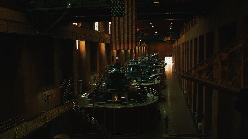 A dimly lit power plant hall with a row of large generator units beneath a hanging American flag