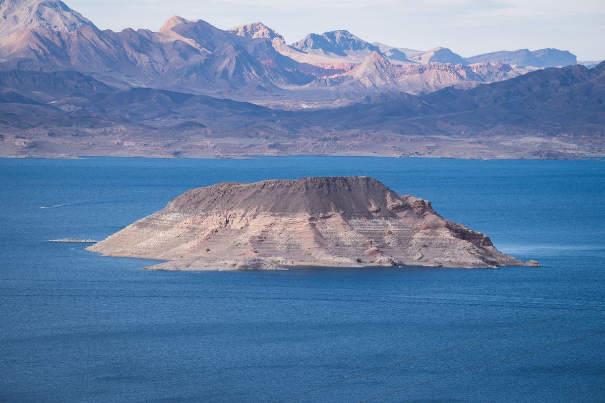 A rocky island with pale mineral bands sits in blue lake water beneath rugged desert mountains