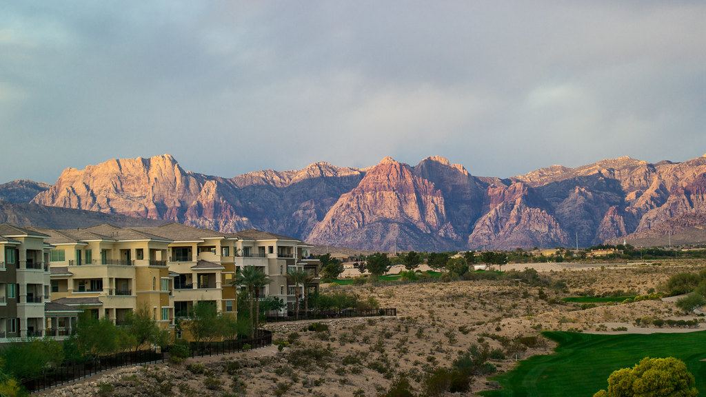 Multistory residential buildings and desert scrub beneath red rock mountains under a cloudy gray sky
