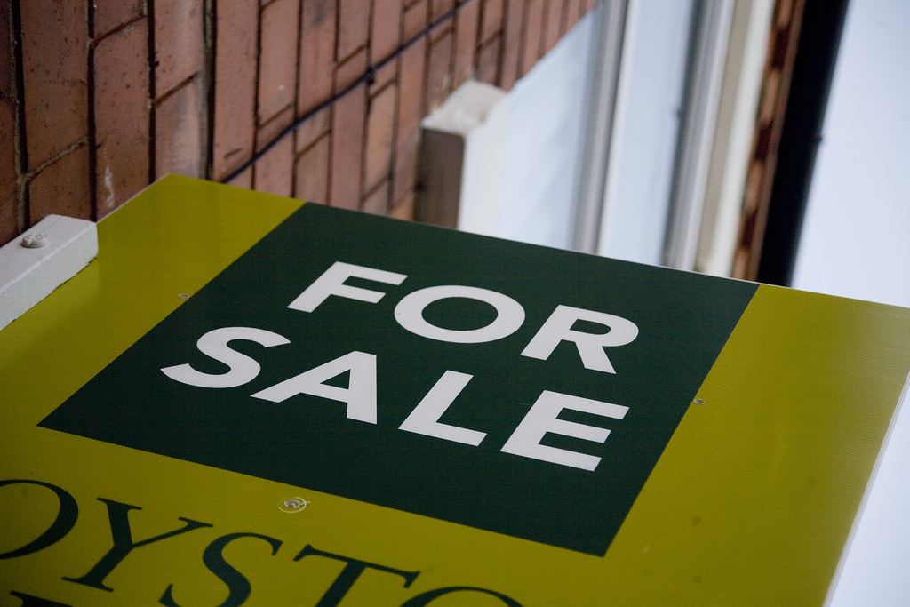 A green and yellow 'For Sale' sign mounted on a brick building beside a window