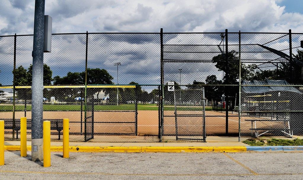 Empty softball field with a dirt infield seen through a chain-link fence under a cloudy sky