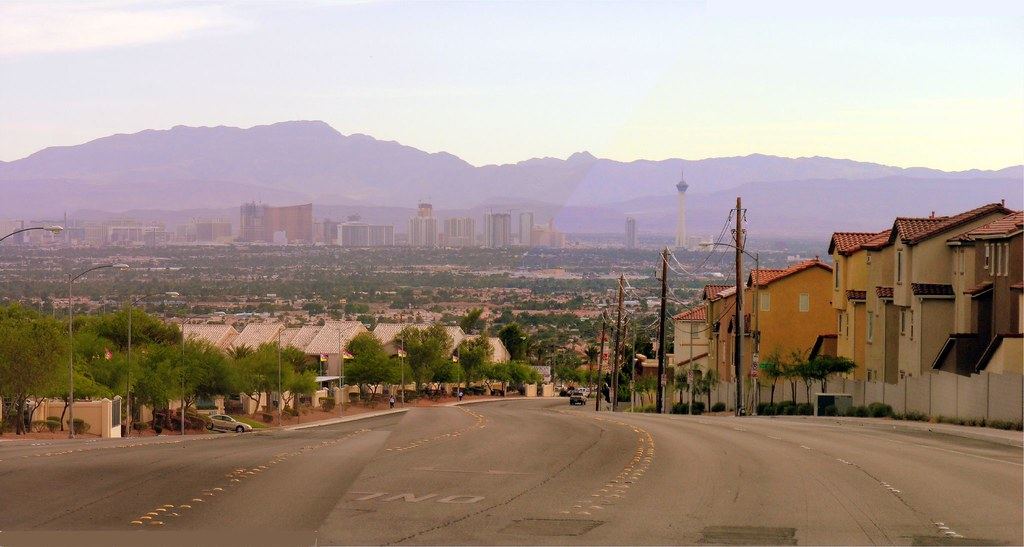 A wide road through a residential neighborhood leads toward the Las Vegas skyline and hazy mountains