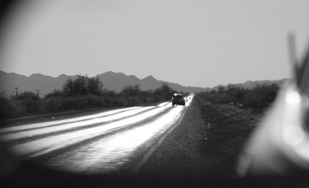 Black-and-white view of a sunlit desert highway with a vehicle, scrub brush and distant mountains